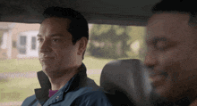 a man in a blue jacket sits in the back seat of a car with another man