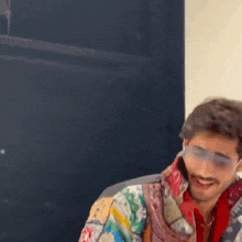 a man wearing sunglasses and a colorful jacket is smiling while sitting in a chair .