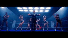 a man in a tuxedo is standing in a boxing ring surrounded by female dancers
