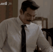 a man with a mustache wearing a white shirt and tie is sitting at a table in front of an ant logo