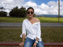 a woman wearing sunglasses sits on a bench in front of a grassy field
