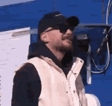 a man wearing sunglasses and a hat is standing in front of a blue wall and talking on a cell phone .