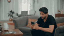 a man in a black shirt is sitting on a couch looking at his phone