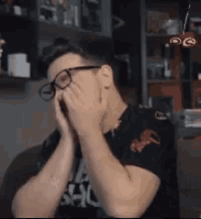 a man wearing glasses is covering his face with his hands while sitting in a chair .