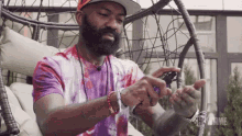 a man with a beard wearing a tie dye shirt is sitting in a wicker chair looking at his watch .