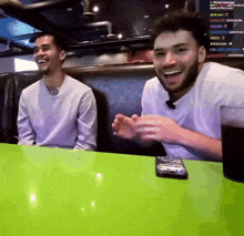 two men are sitting at a table with a green table cloth and a remote control on it