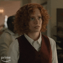 a woman with red curly hair is wearing a vest and white shirt .