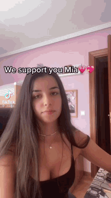 a woman in a black tank top is standing in front of a pink wall with the words we support you mia .