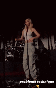 a woman in a white tank top and gray pants is dancing on a stage in front of a drum set ..