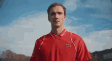 a man wearing a red shirt with a lacoste logo on it is standing in front of a blue sky .