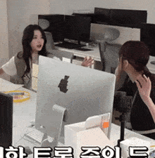 two women are sitting at a desk in front of an apple computer monitor