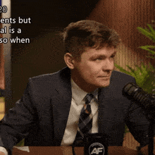 a man in a suit and tie sitting in front of a microphone with a mug that says af on it