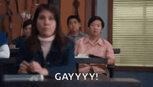a woman is sitting at a desk in a classroom with other people and making a funny face .