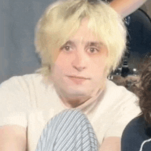 a man with blonde hair is sitting down and looking at the camera with a serious look on his face .