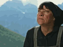 a woman wearing a black hat and overalls looks up at the sky with mountains in the background