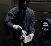 a man in a hooded jacket is pouring a cup of coffee to another man in a chair .