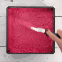 a person is using a spatula to spread red frosting on a square pan