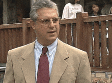 a man in a tan suit and red tie stands in front of a fence