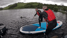 a man in a red life jacket is helping another man on a surfboard