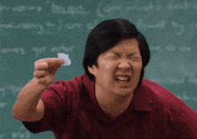 a man in a red shirt is making a funny face in front of a blackboard that says ' economics ' on it