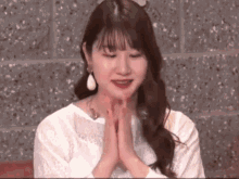 a woman wearing a white shirt and earrings is sitting with her hands folded in front of her face .