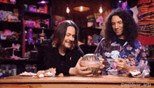 two men are sitting at a table and one is holding a watermelon with spaghetti on it