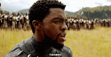 a man in a black suit is standing in a field with a crowd of people behind him and says yibambo .