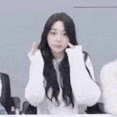 a woman with long black hair is wearing a white sweater and making a heart shape with her fingers
