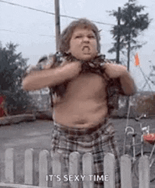 a fat boy is standing in front of a fence with his shirt off .