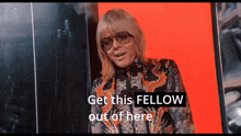 a woman wearing sunglasses stands in front of a red background and says get this fellow out of here