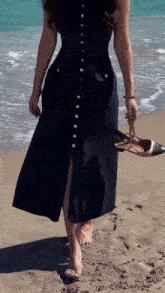 a woman in a black dress is standing on the beach holding her shoes