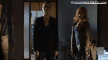 two women standing next to each other with the website shadowhunterstv.com in the background