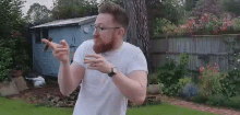 a man with a beard and glasses is standing in a garden holding a cigarette .