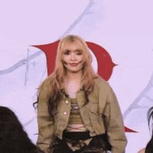 a woman with blonde hair and a green jacket is standing in front of a red letter d