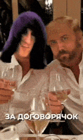 two men are sitting at a table holding wine glasses and one of them has a purple hat on his head