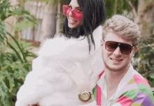 a man wearing sunglasses and a watch stands next to a woman wearing pink sunglasses