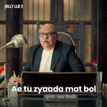 a man in a suit sits at a desk with the words " ae tu zyaada mat bol " above him
