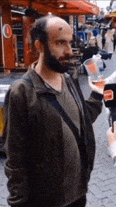 a man with a beard and a bald head is standing on a sidewalk holding a glass of beer .