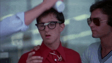 a man wearing glasses and a red shirt is being helped by a man wearing sunglasses