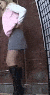 a woman in a pink shirt and gray skirt is standing in front of a brick wall .