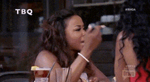 a woman is sitting at a table with a drink in her hand and another woman is applying makeup to her face .