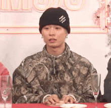 a man wearing a black beanie that says miami loves you sits at a table with wine glasses
