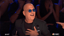 a bald man wearing sunglasses and a black suit laughs while being pointed at