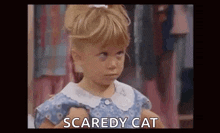 a little girl in a blue dress is holding a doll and saying scaredy cat .