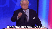 a man in a suit and tie stands at a podium with the words " and the dream lives on " behind him