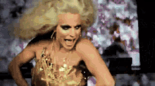 a drag queen with blonde hair and a gold dress is dancing .