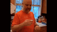 a bald man in an orange shirt is holding a cup in his hand .