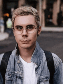 a young man wearing glasses and a denim jacket is looking at the camera