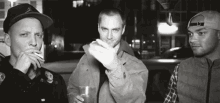 three men are standing next to each other in a black and white photo . one of the men is smoking a cigarette .