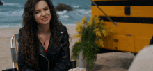 a woman in a leather jacket is sitting in a chair on the beach in front of a yellow school bus .